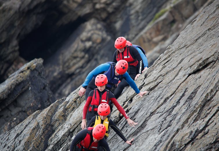 chlidren in red helmets and wetsuits climb down a cliff