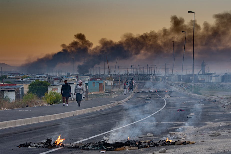 People walk past shacks where burning material blocks the road and smokes rises in the background