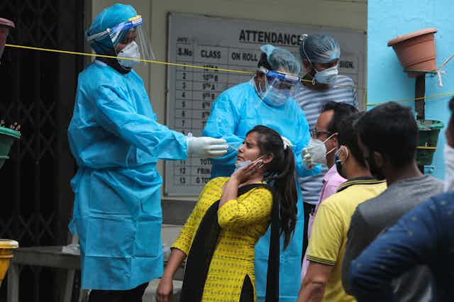 Indian woman leans back as health care worker swabs her nose for COVID-19 test, there is a line of people behind her