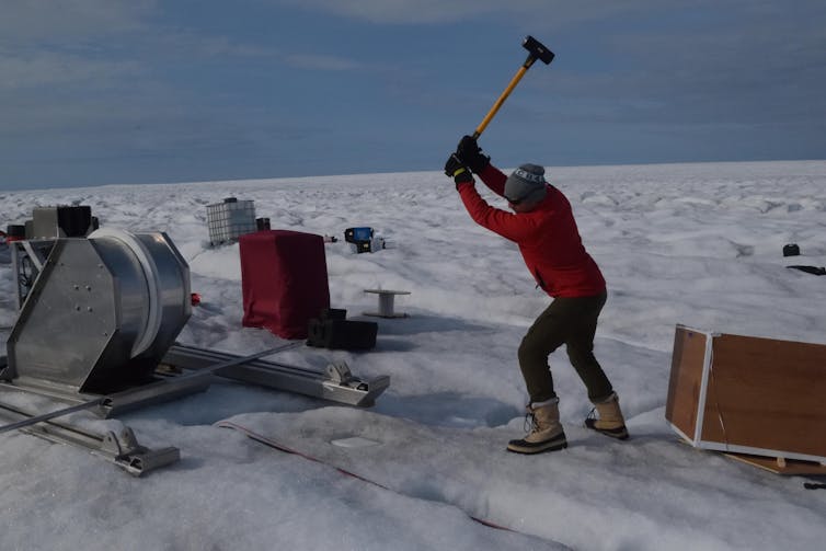 A man raises a hammer above an icy tundra surrounded by equipment