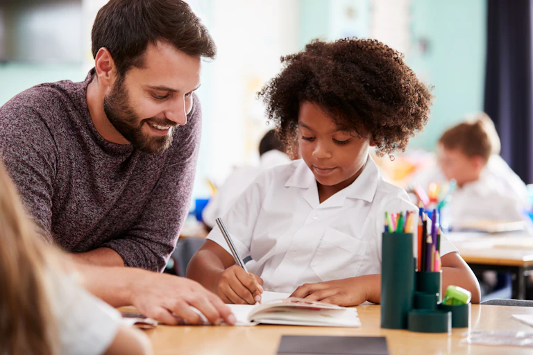 Male teacher helping student with something at her desk.