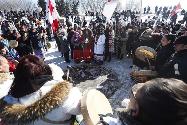 Indigenous protesters, some wearing ribbon skits, some holding drums stand in a circle in the winter.