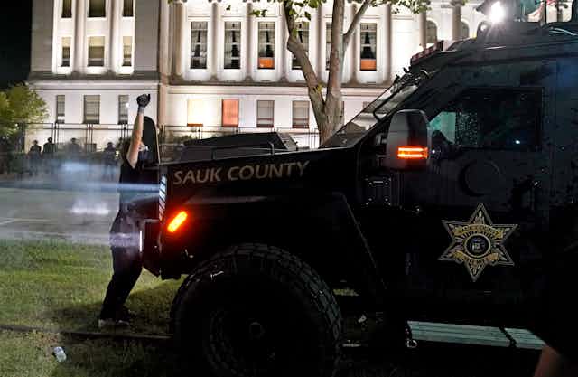 An armored vehicle with a sheriff's badge logo on the door moves toward a masked protester