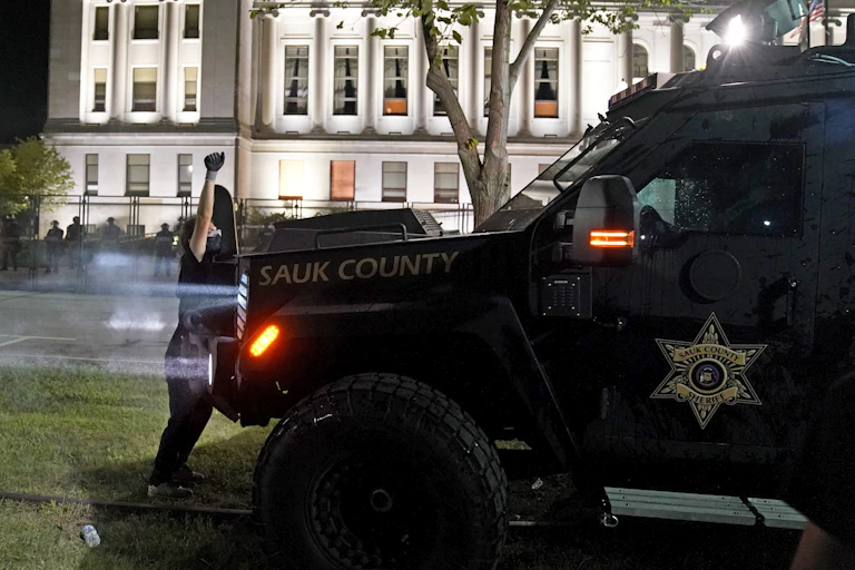 An armored vehicle with a sheriff's badge logo on the door moves toward a masked protester