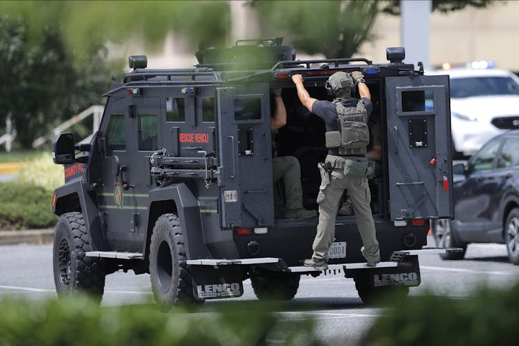 An armored vehicle drives with a helmeted man holding onto the back