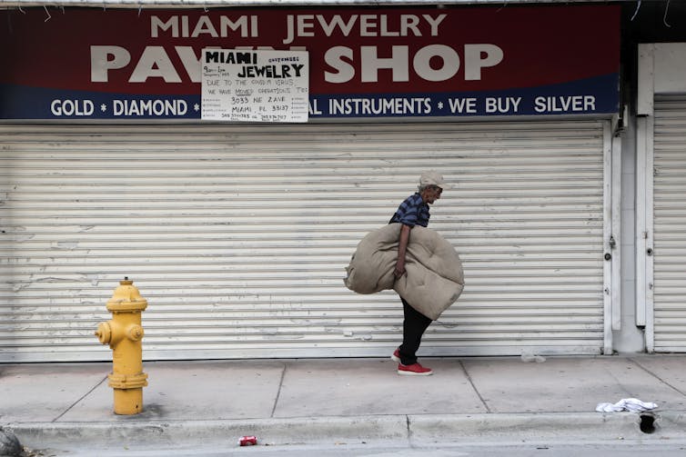 an older man holding a large cloth bundle under his arm walks along the sidewalk in front of the metal grate of a closed pawn shop