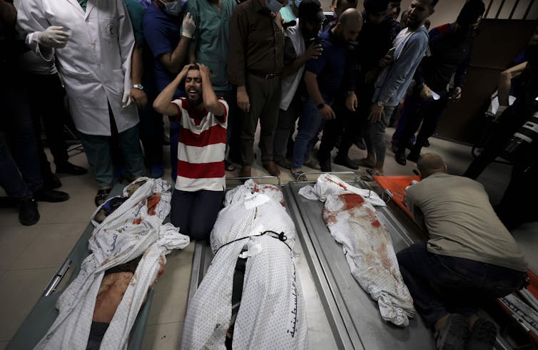 A man in grief on his knees next to the bodies of three of his family members laid out on stretchers.