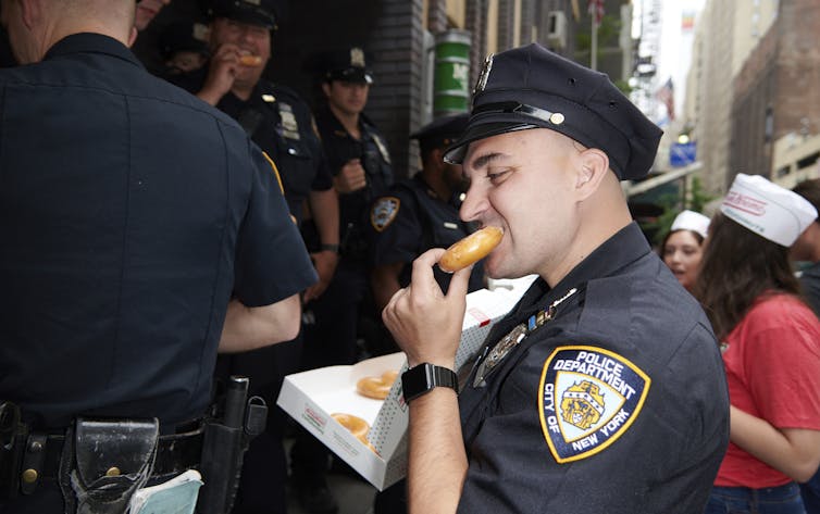A New York police office bites into a Krispy Kreme doughnut while holding a box of doughnuts as other officers and Krispy Kreme employees mingle in background