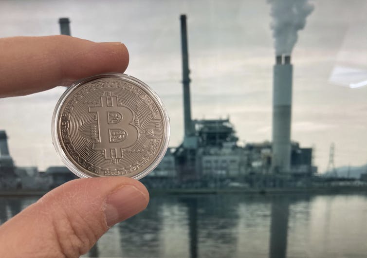 fingers hold bitcoin in front of smokestacks