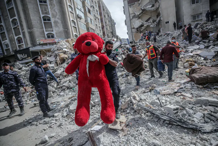 A Palestinian man holds up a large red teddy bear from the ruins of an apartment block.