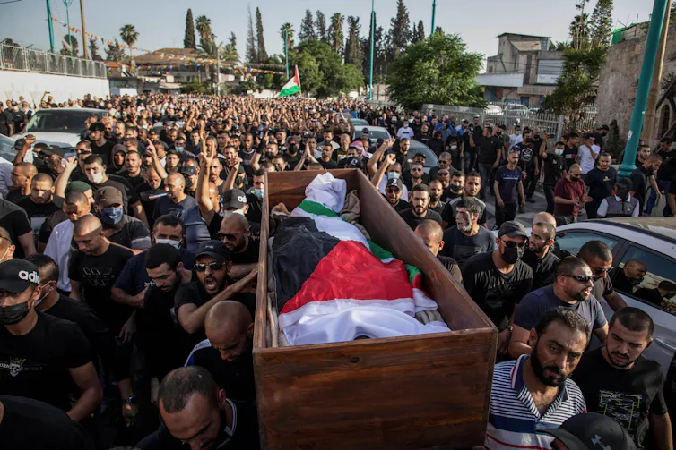 Arab Israeli men carry the body of a man wrapped in a Palestinian flag surrounded by crowds of people at a funeral.