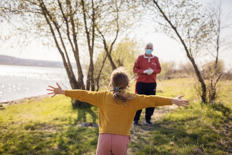 A young girl runs to hug her grandpa, who is wearing a mask outdoors.