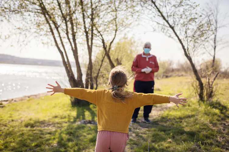 A young girl runs to hug her grandpa, who is wearing a mask outdoors.