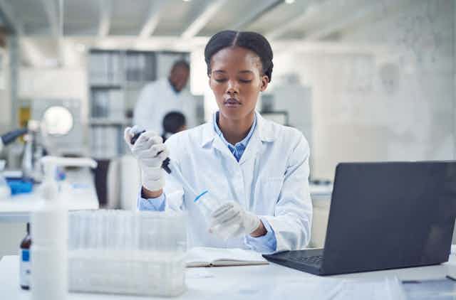 Woman in white laboratory coat and protective gloves handles equipment, with a computer on the desk