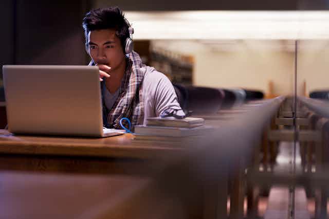 Un jeune étudiant portant des écouteurs suit un cours en ligne devant son ordinateur portable dans une bibliothèque