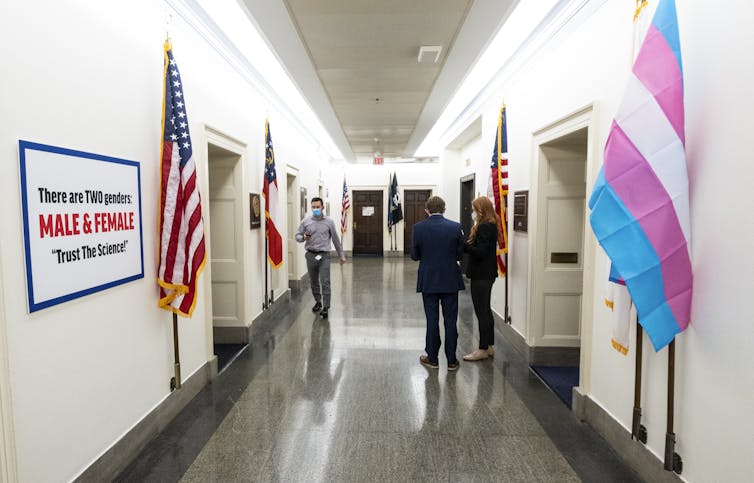 A transgender rights flag stands across from a transphobic sign in hallway of government offices