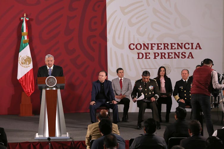 O presidente do México fala em um púlpito em um palco com uma pequena multidão de funcionários do governo sentados nas proximidades