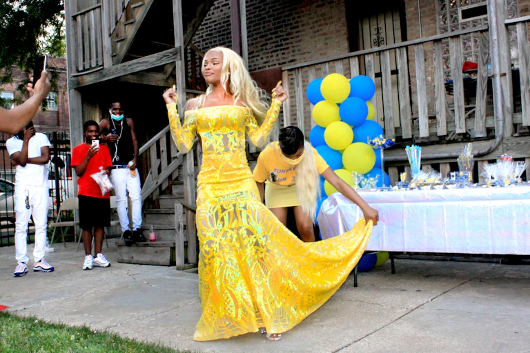 A young Black woman wears a yellow dress as a group of her siblings take photos of her.