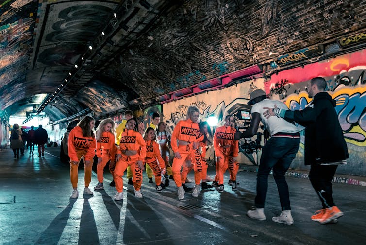 Dancers in matching orange tracksuits perform for a music video in a graffiti-covered tunnel in London
