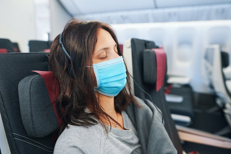 A woman sleeping on a plane, wearing headphones and a face mask.
