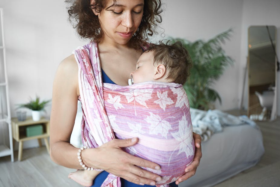 Woman holds baby close to her chest, who is sleeping wrapped in a soft fabric sling.