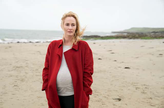Pregnant woman in a red coat walks on a beach.