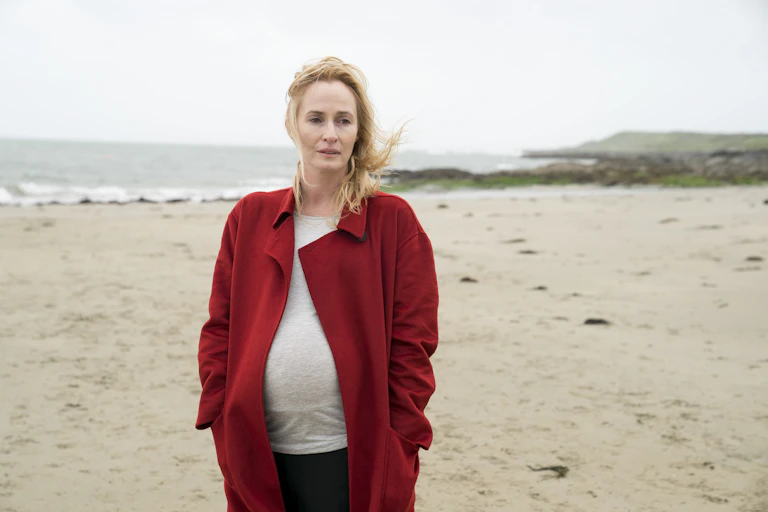 Pregnant woman in a red coat walks on a beach.