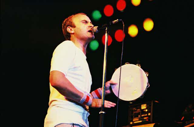 Phil Collins singing and plating a tambourine on stage.