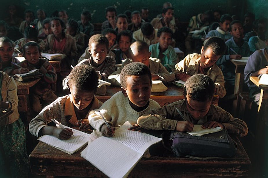 Children sitting at desks in a crowded classroom, poorly lit