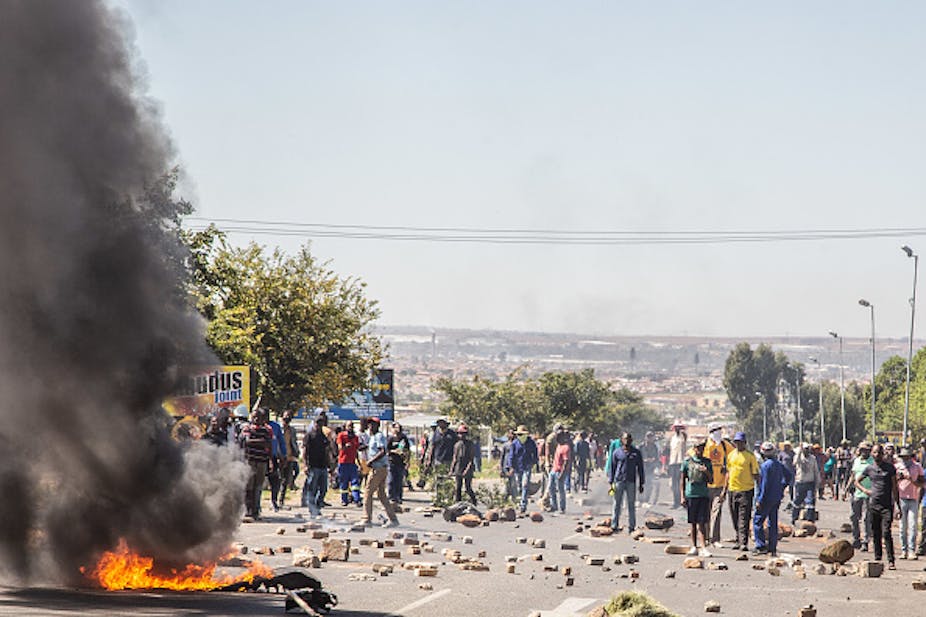 Lots of rocks and a burning tyre litter a tarred road where protesters are standing