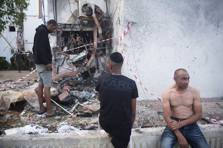 Three people looking at rubble from a building destroyed by a rocket attack.