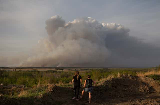Two people watch a large cloud of smoke rising over fields. 