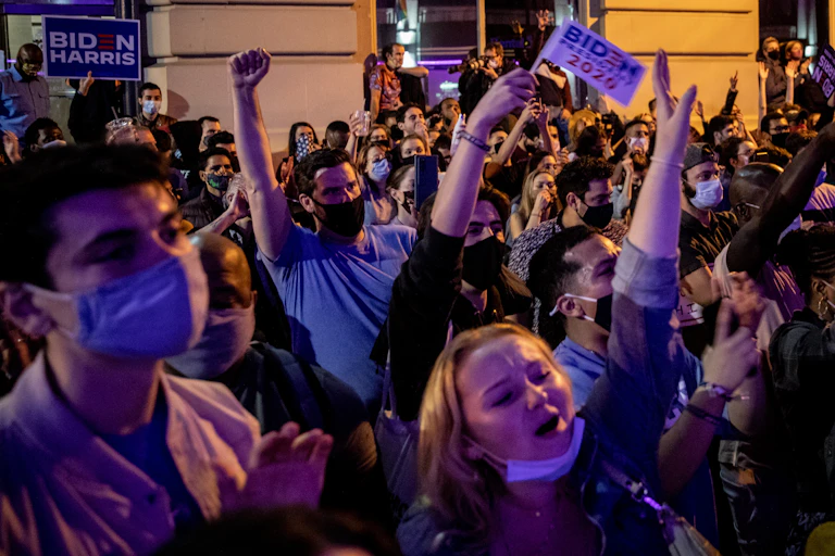 Many people celebrating outdoors in the street, with Biden-Harris signs.