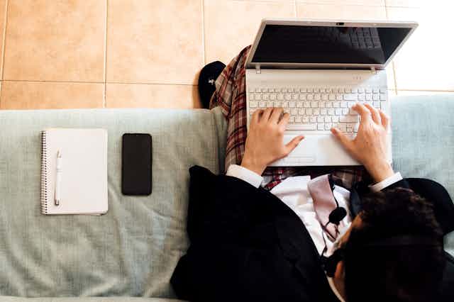 Man wearing a suit jacket and tie but pajama pants types on keyboard of laptop while sitting on couch with a notebook and smartphone next to him
