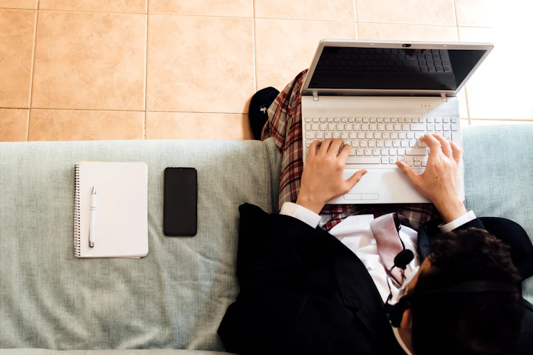 Man wearing a suit jacket and tie but pajama pants types on keyboard of laptop while sitting on couch with a notebook and smartphone next to him