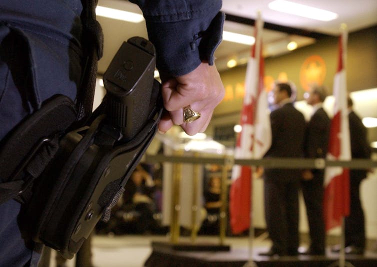 A police officer's hand rests on his gun as politicians speak in the background.