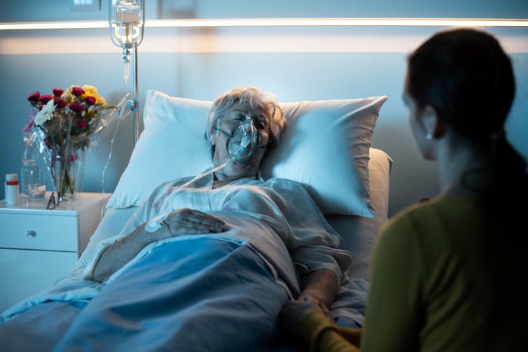 A woman wearing an oxygen mask lies in a hospital bed. Another woman is comforting her.