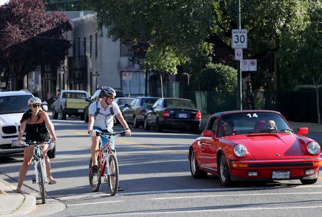 Two cyclists wait at a traffic light alongside a red sports car