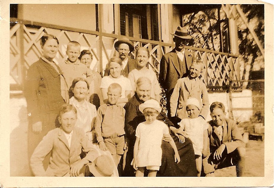 A sepia-toned photograph shows a group of men, women and children in older clothing outside a home