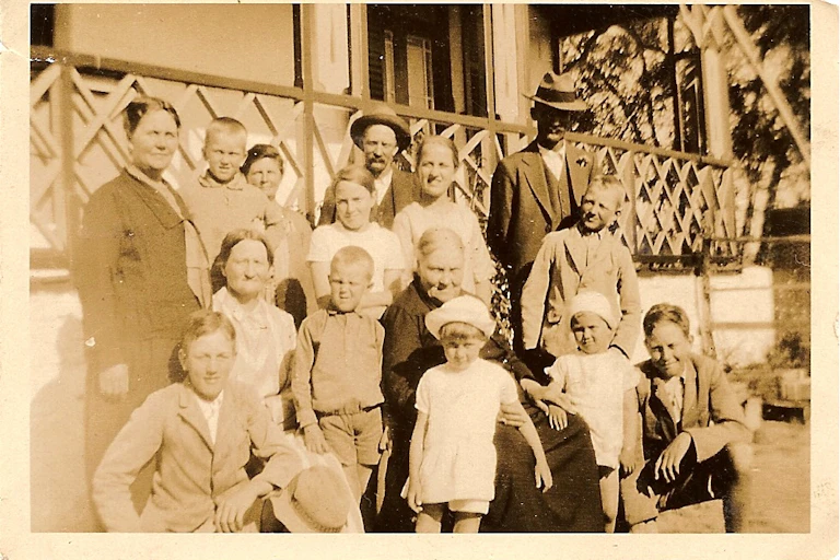 A sepia-toned photograph shows a group of men, women and children in older clothing outside a home