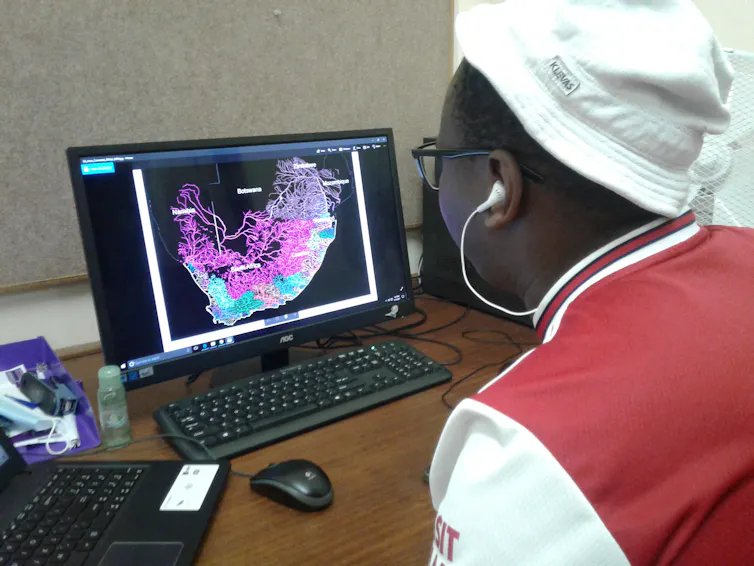 A person wearing a white hat and a red and white shirt, with white headphones, looks at a computer screen.