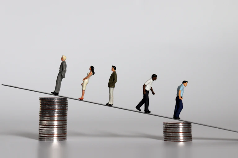 Small human model figures walk a see-saw between two different piles of coins