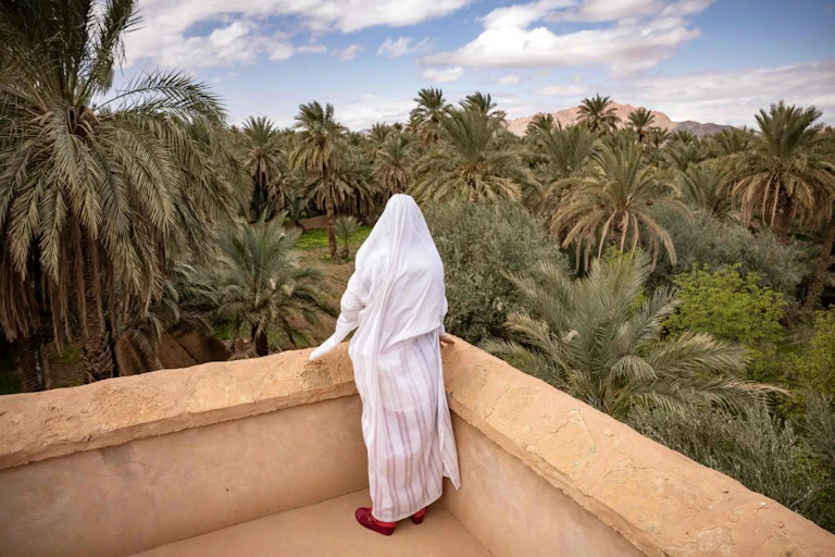 A woman in a head covering and robe stands at a viewpoint overlooking some palm trees.