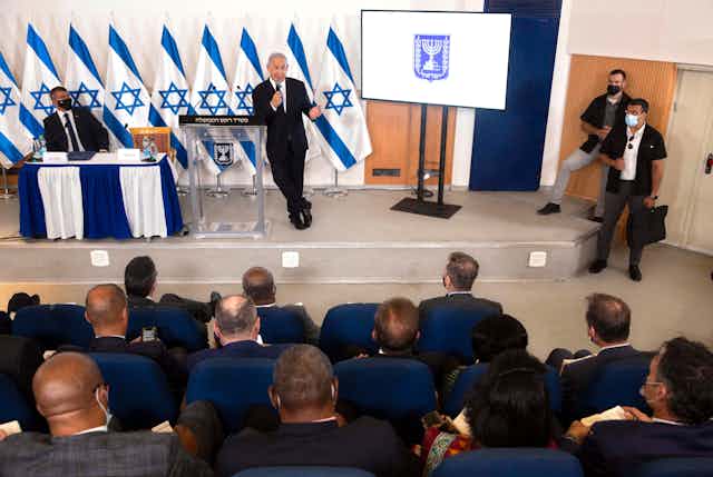 Israeli Prime Minister Benjamin Netanyahu, standing in front of multiple Israeli flags, giving a briefing to foreign diplomats from a military base in Tel Aviv.