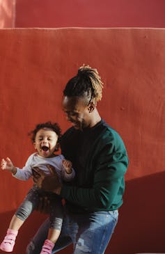 A father in a green top holds a toddler in front of a red wall