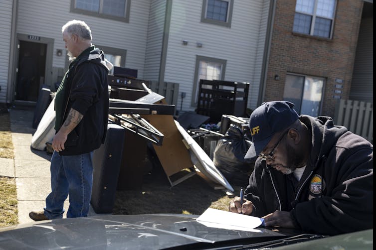 Man leans on hood of car to sign document. Behind him is another man standing in front of furniture piled on the front lawn of an apartment building