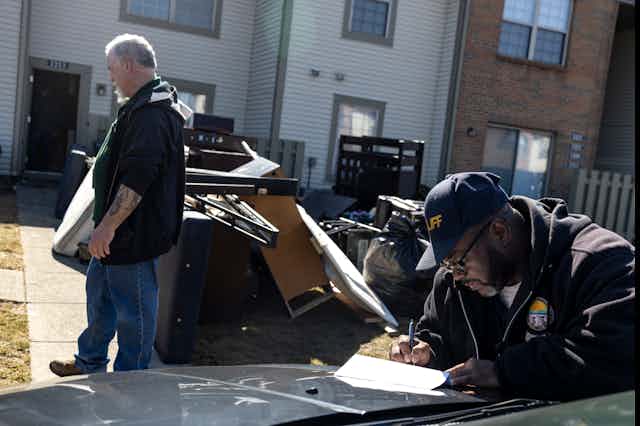A man turns away from a pile of his belongings on a lawn, while a bailiff completes paperwork