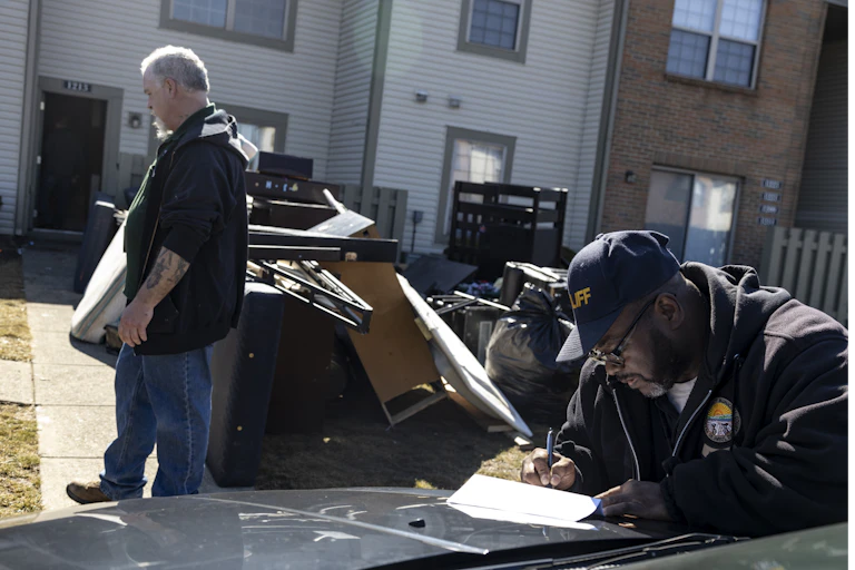 A man turns away from a pile of his belongings on a lawn, while a bailiff completes paperwork