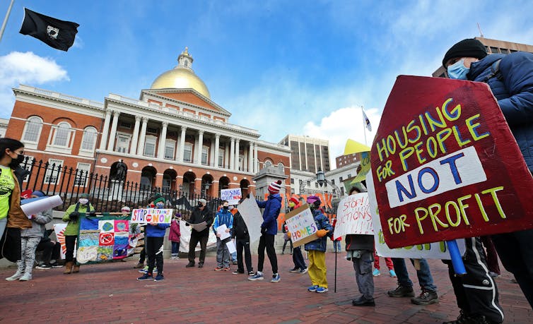 People hold signs in front of a state capitol building. Signs read Housing for People Not for Profit and Stop evictions now