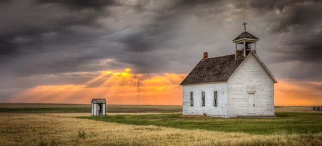 A old church building in a field. Above sunlight peeks through grey clouds.